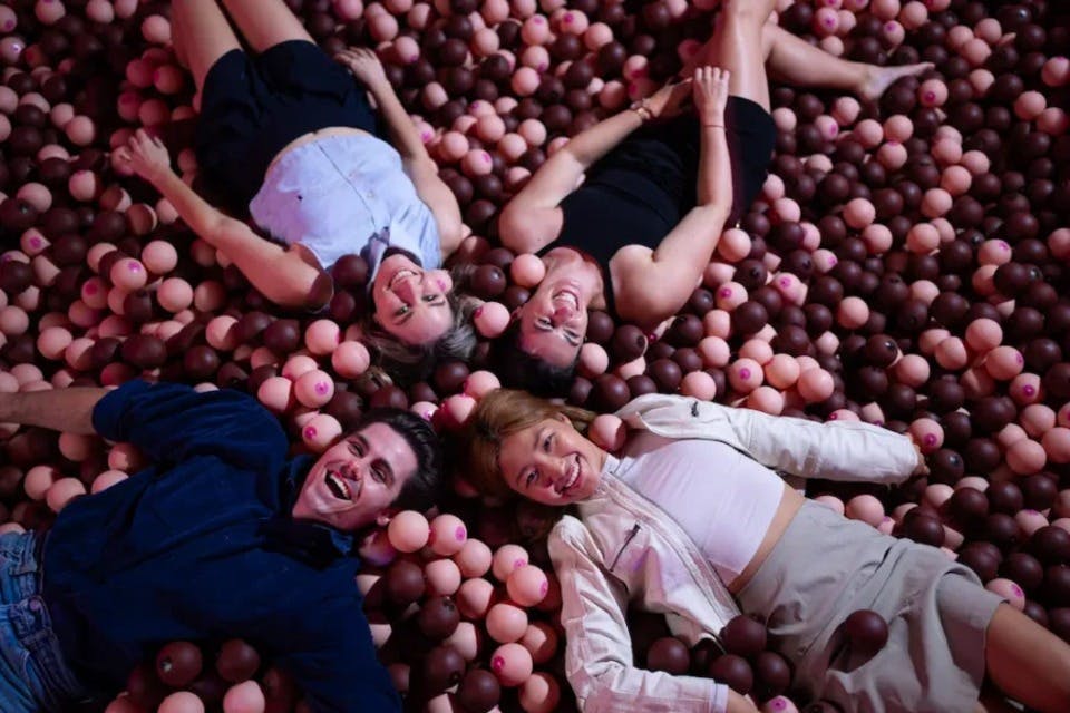 갈색과 분홍색 공이 가득한 공 구덩이에 네 명이 등을 대고 누워 웃고 있습니다.