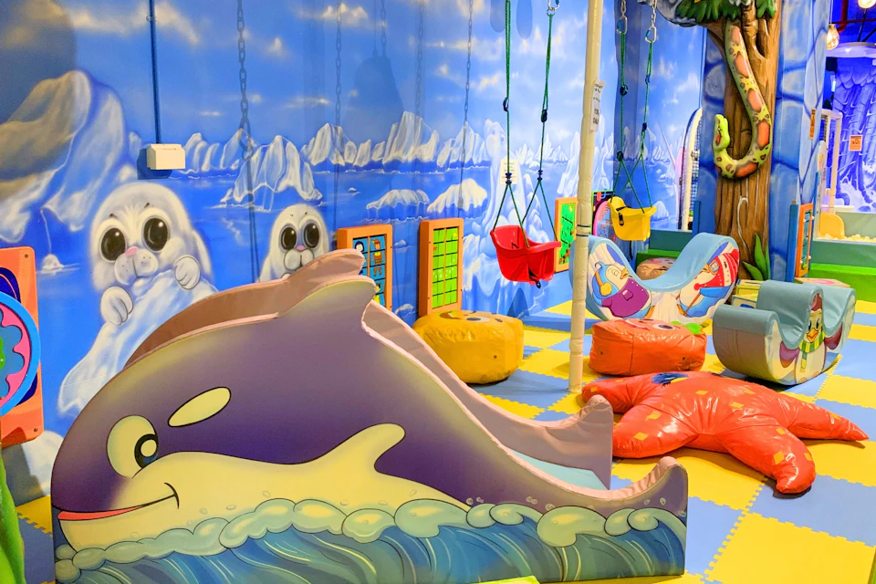 A colorful indoor children's play area with ocean-themed decor, foam slides, swings, and padded seating on a checkered floor.