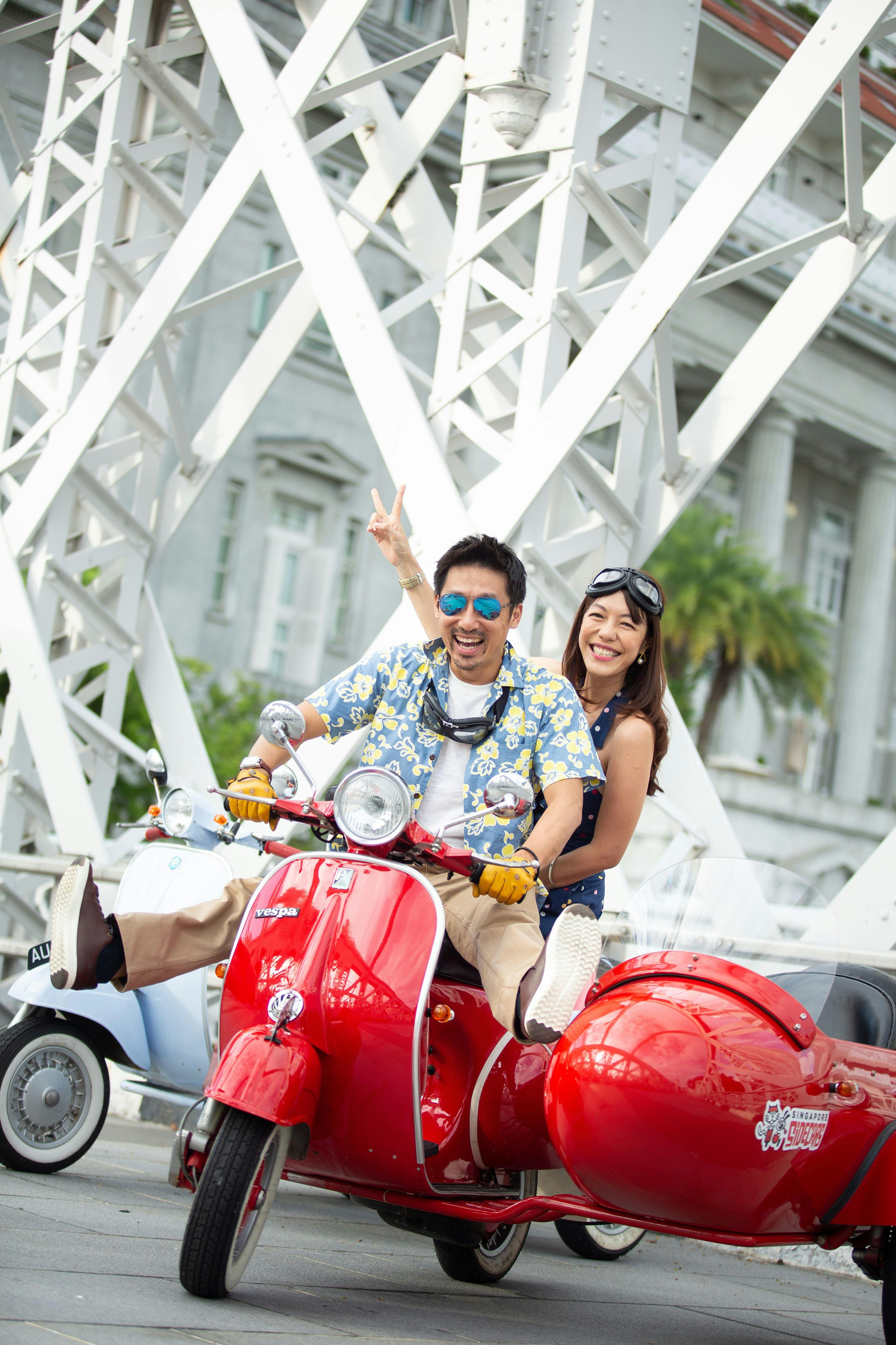 Deux personnes souriant sur une Vespa rouge avec un side-car, sur fond de poutres structurelles blanches et d'un bâtiment.
