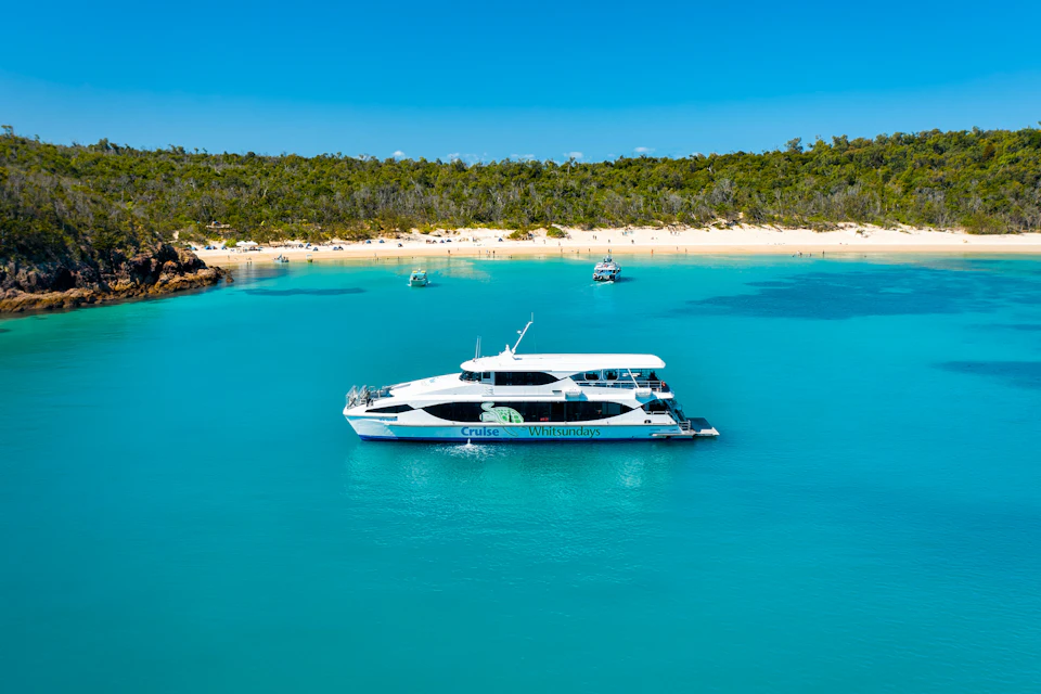 A white and blue catamaran is anchored in clear turquoise water, near a sandy beach with a few boats and lush greenery in the background.