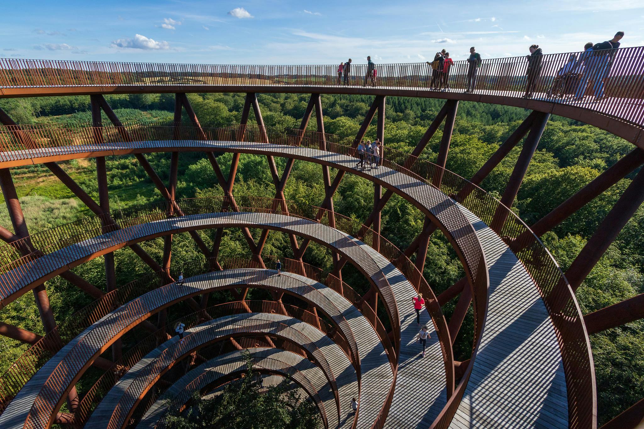 Ludzie idący po spiralnej drewnianej konstrukcji rampy z widokiem na bujny zielony las pod czystym błękitnym niebem.
