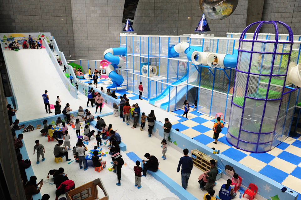 Indoor playground with colorful slides, climbing structures, and children playing. Parents are present, overseeing activities.