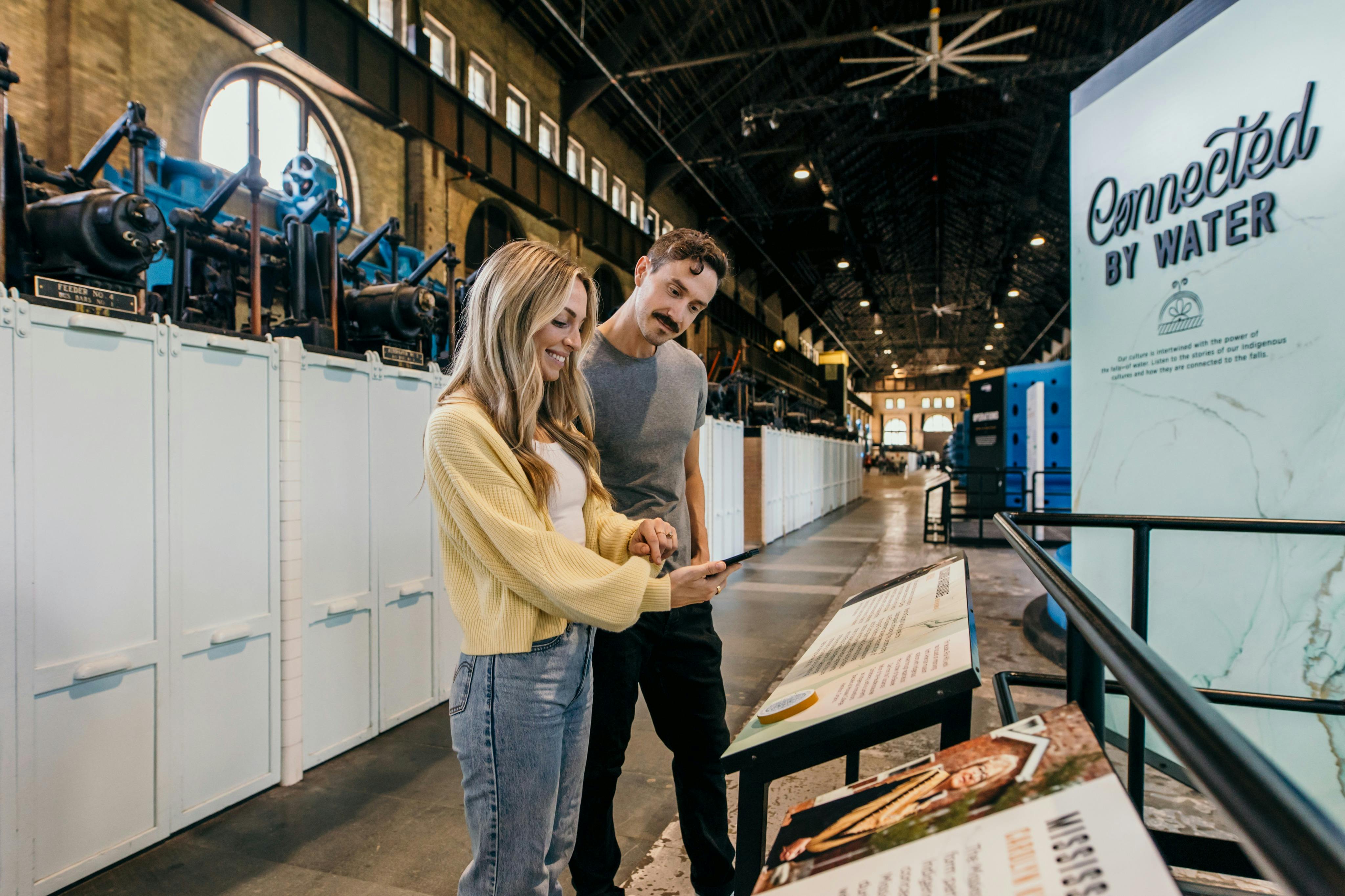 박물관에서 산업 기계와 천장이 높은 방을 배경으로 정보 디스플레이를 읽고 있는 두 사람.