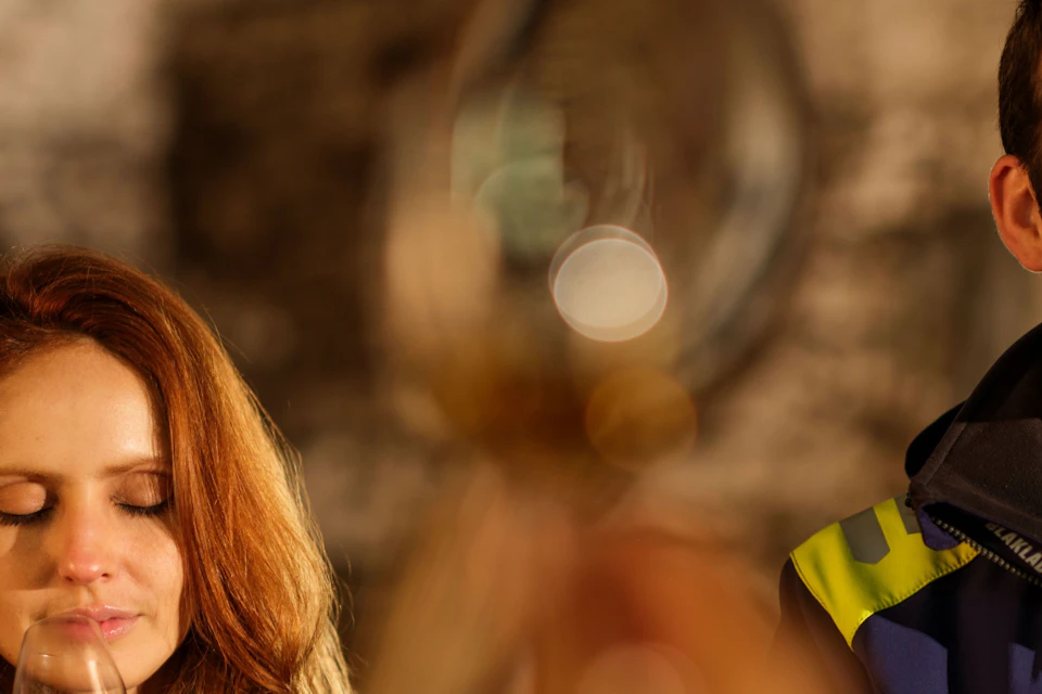 Two people holding glasses with golden liquid, appearing to engage in a tasting session in a warmly lit environment.