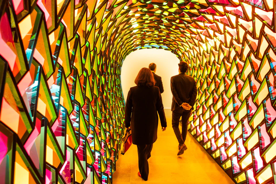 Three people walking through a colorful, illuminated tunnel made of geometric, stained-glass-like panels.