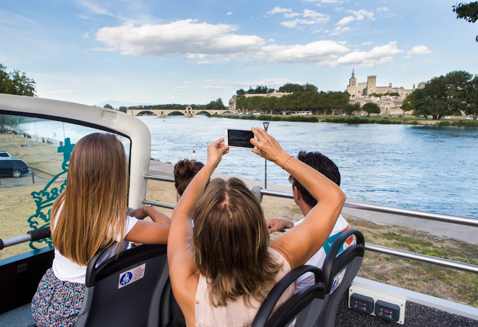 Three people on an open-top bus take a photo of a historic bridge and river under a blue sky with clouds.