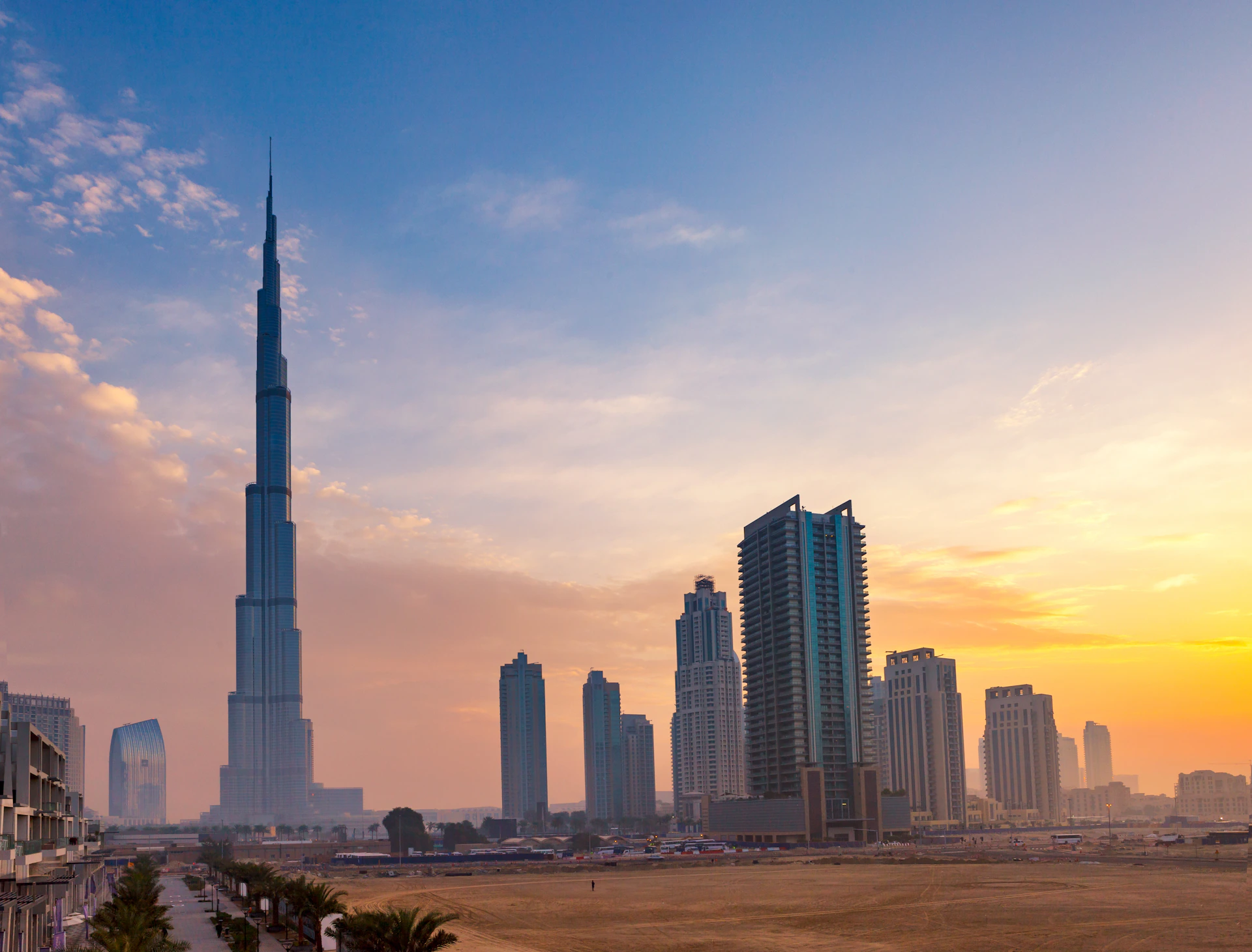 Tall skyscrapers, including the Burj Khalifa, against a vibrant sunset sky in a cityscape with some palm trees and sandy ground in the foreground.