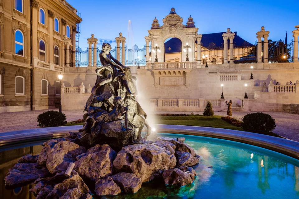 A decorative fountain with sculptures in front of an ornate building and gate, illuminated at twilight.