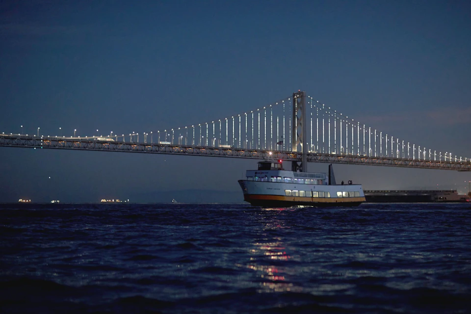 A ferry boat on a dark waterway at night with a lit suspension bridge in the background.