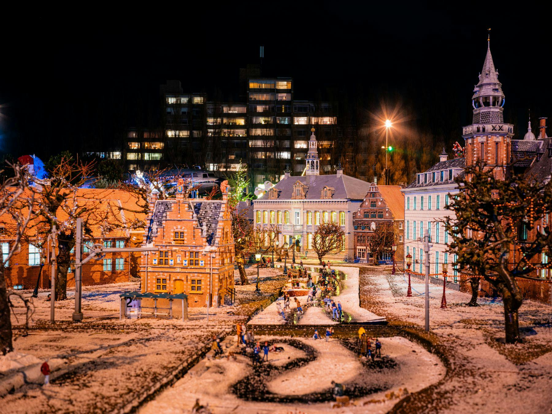 街の明かりを背景に、夜の雪に覆われたミニチュアの村のシーンでは、照らされた建物、木々、小道に小さな人物が描かれています。