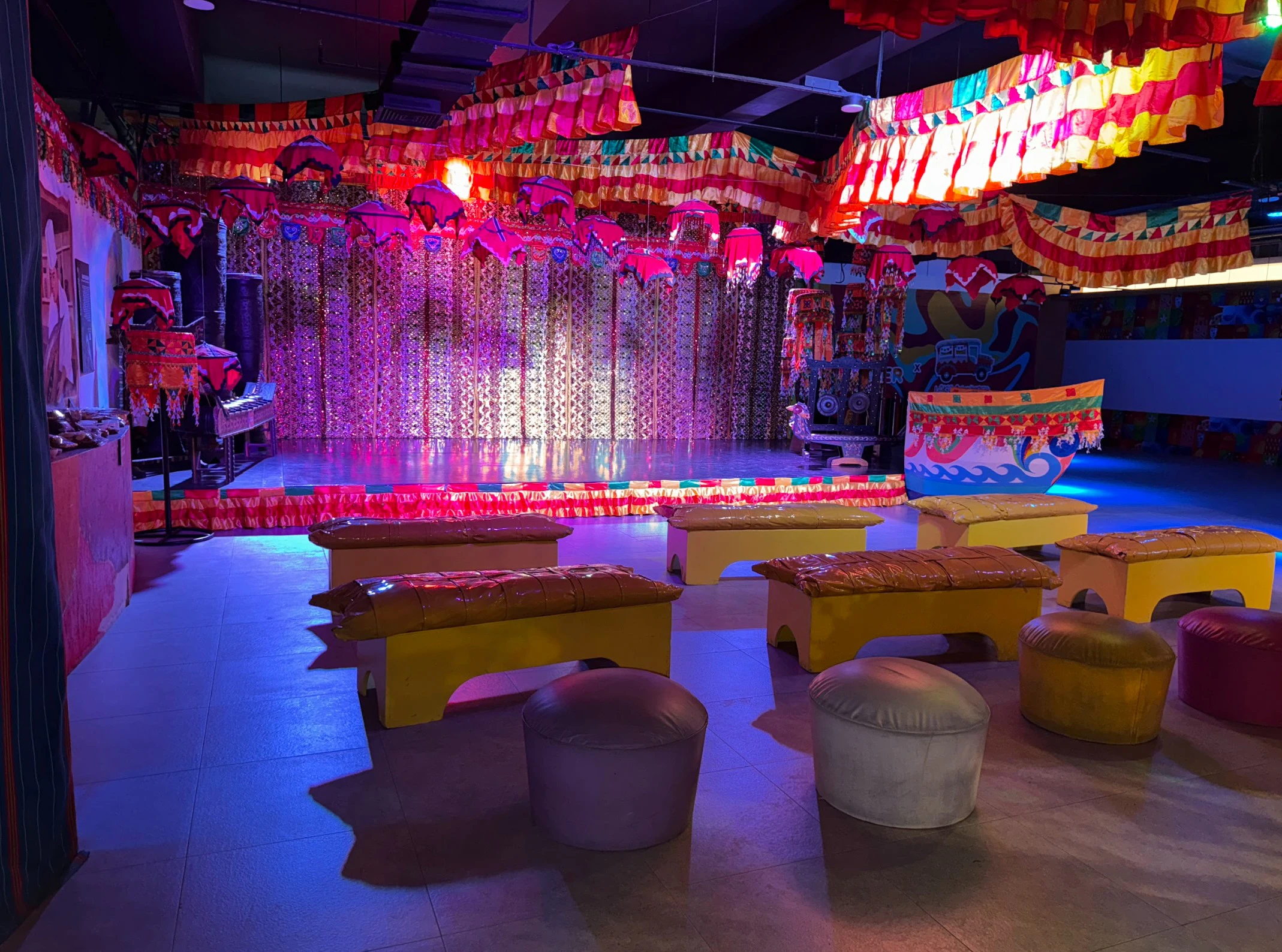 A colorful indoor stage setup with vibrant fabric decorations, benches, and stools arranged in front of the stage.