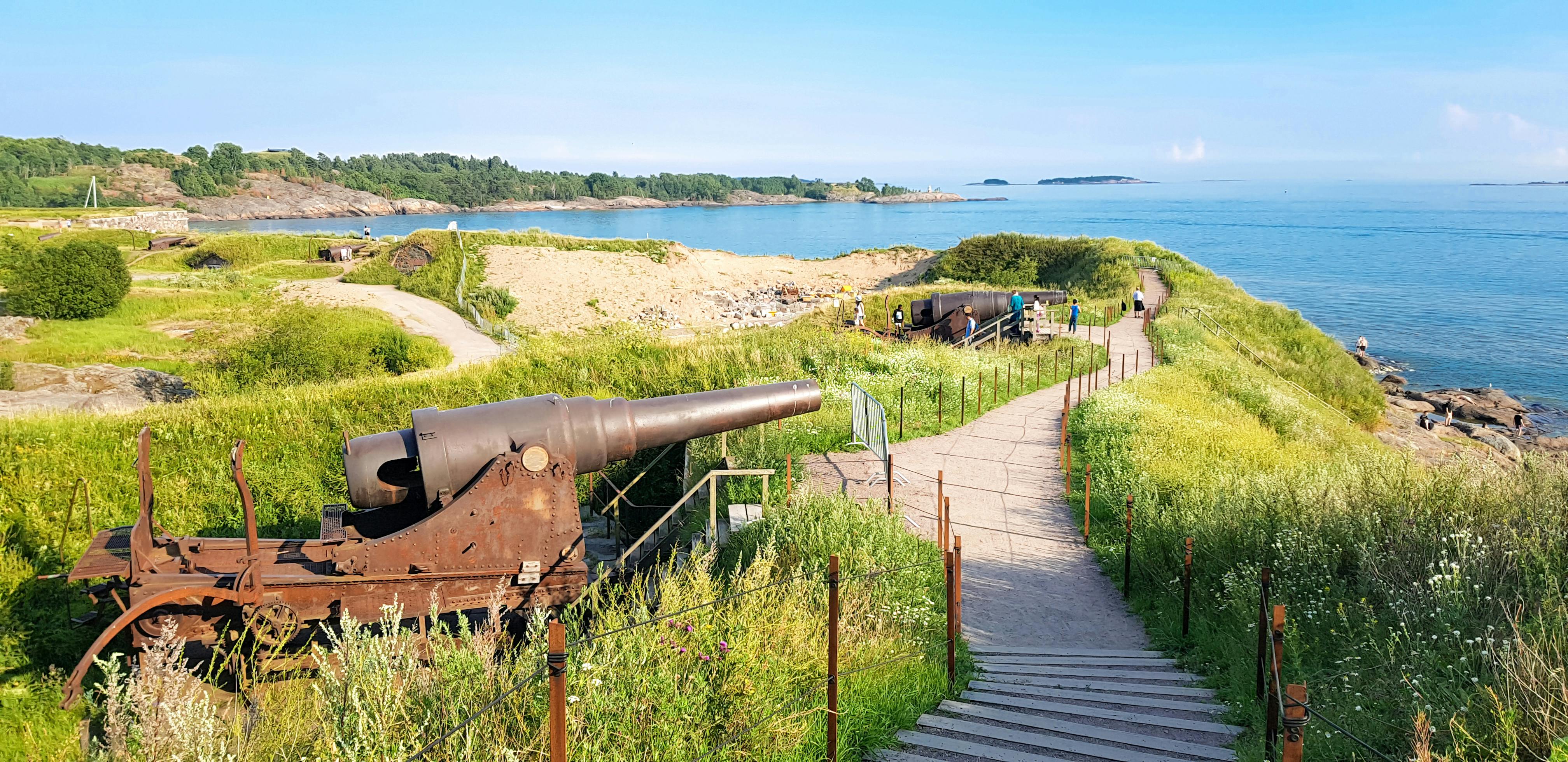 Cannoni arrugginiti che si affacciano su un paesaggio costiero panoramico con sentieri, vegetazione e qualche persona che passeggia.