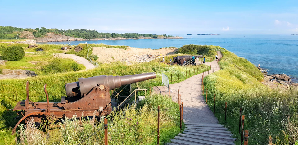 Rusty cannons overlooking a scenic coastal landscape with paths, green vegetation, and a few people walking.