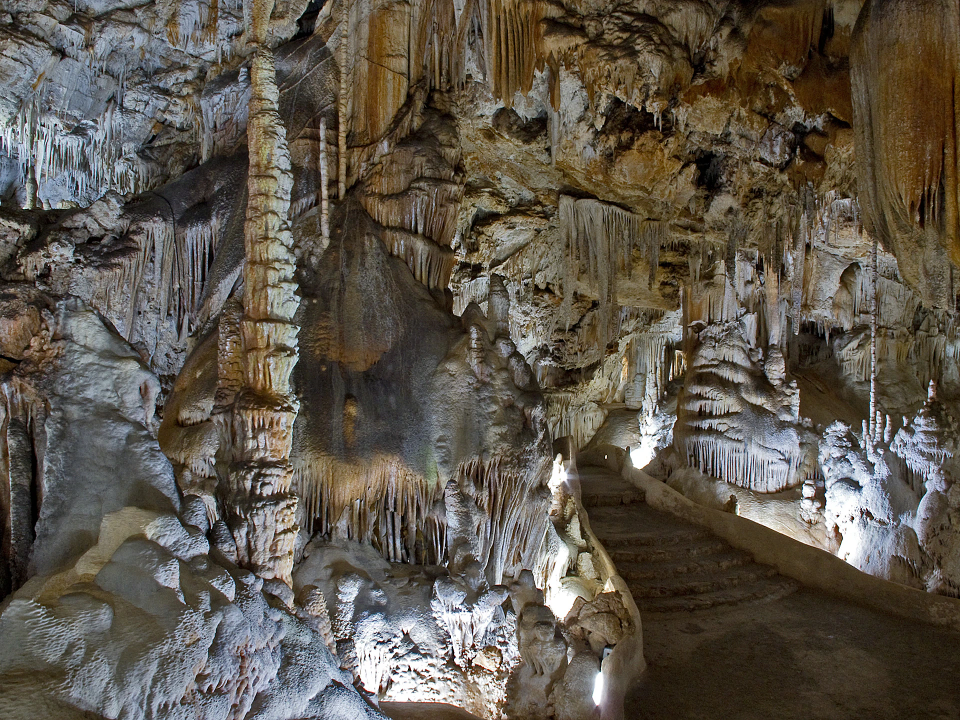 A cave interior featuring numerous stalactites and stalagmites with a lit pathway and stone steps leading deeper into the cave.