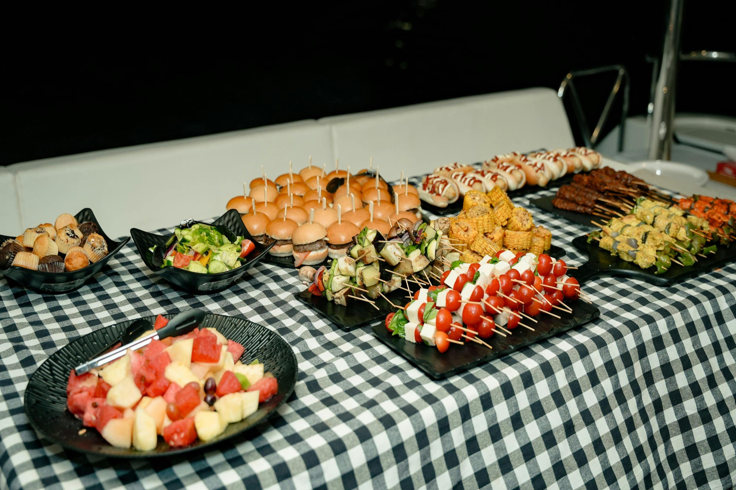 Ein kariertes Tischtuch, auf dem in Schalen und Tabletts Schieber, Spieße, Maiskolben, Obst und Salate angeordnet sind.