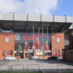 Utsidan av en tegelstadion med stora väggmålningar, flaggor och bilar parkerade framför. Texten lyder: "VI KOMMER INTE för att LEKA."