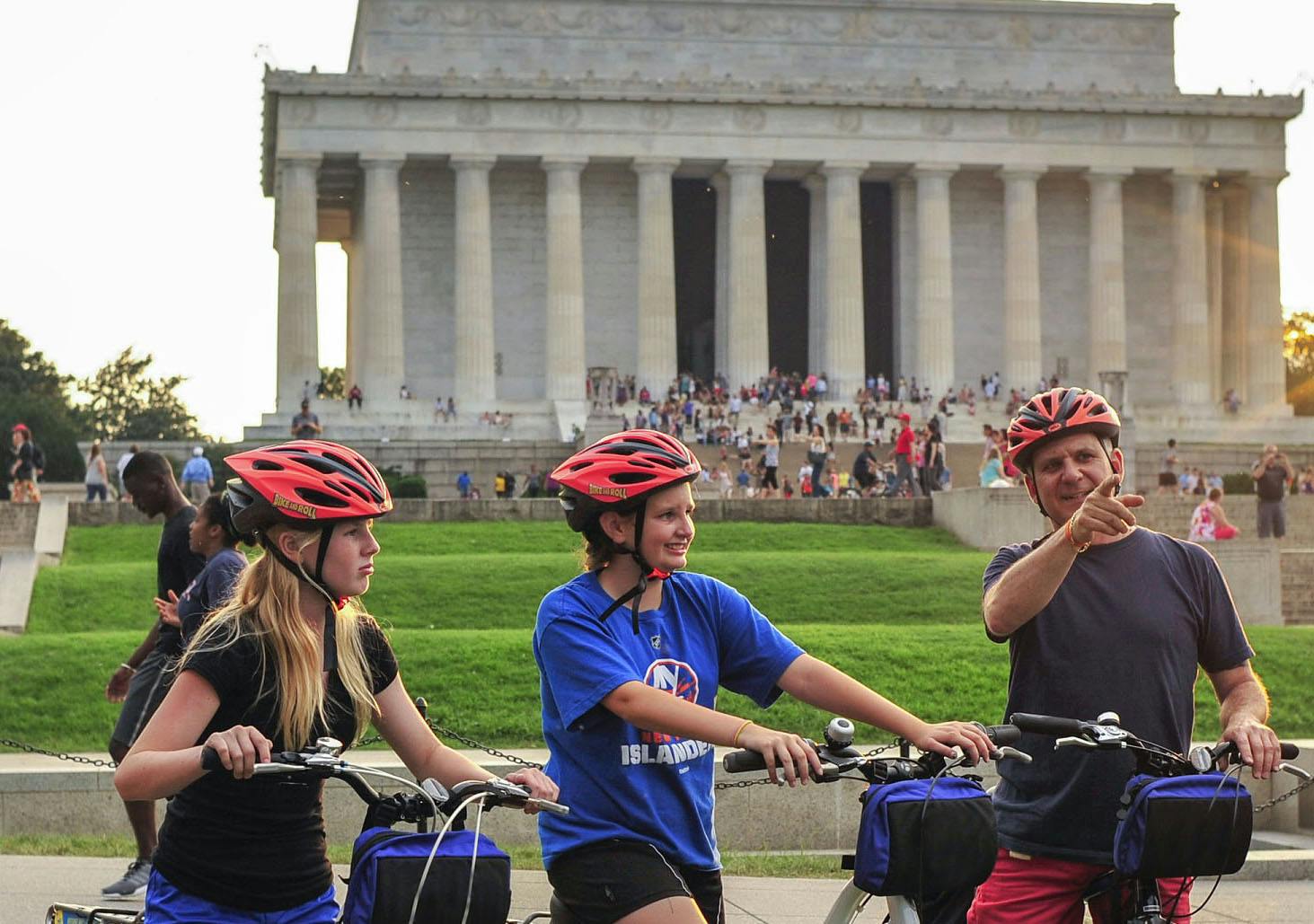 Two people wearing helmets stand with bicycles, smiling and pointing, in front of the Lincoln Memorial.