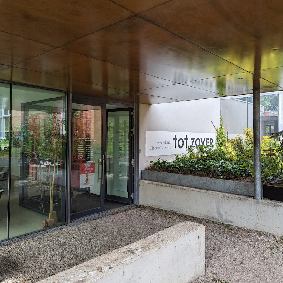 Entrance to a modern building with glass doors, a partial view of an office inside, and a sign reading "tot ZOVER" on the wall outside.