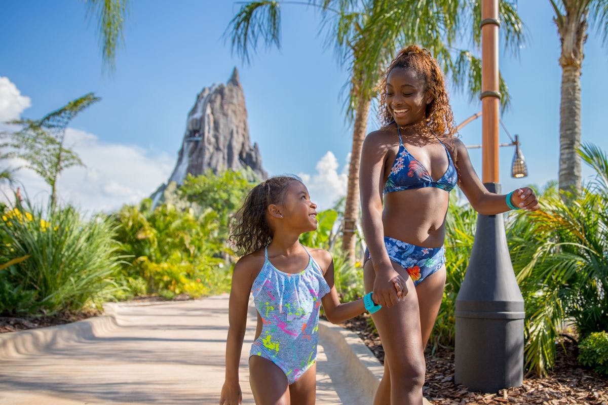 Due ragazze sorridenti in costume da bagno camminano mano nella mano su un sentiero tropicale con palme e una montagna sullo sfondo.