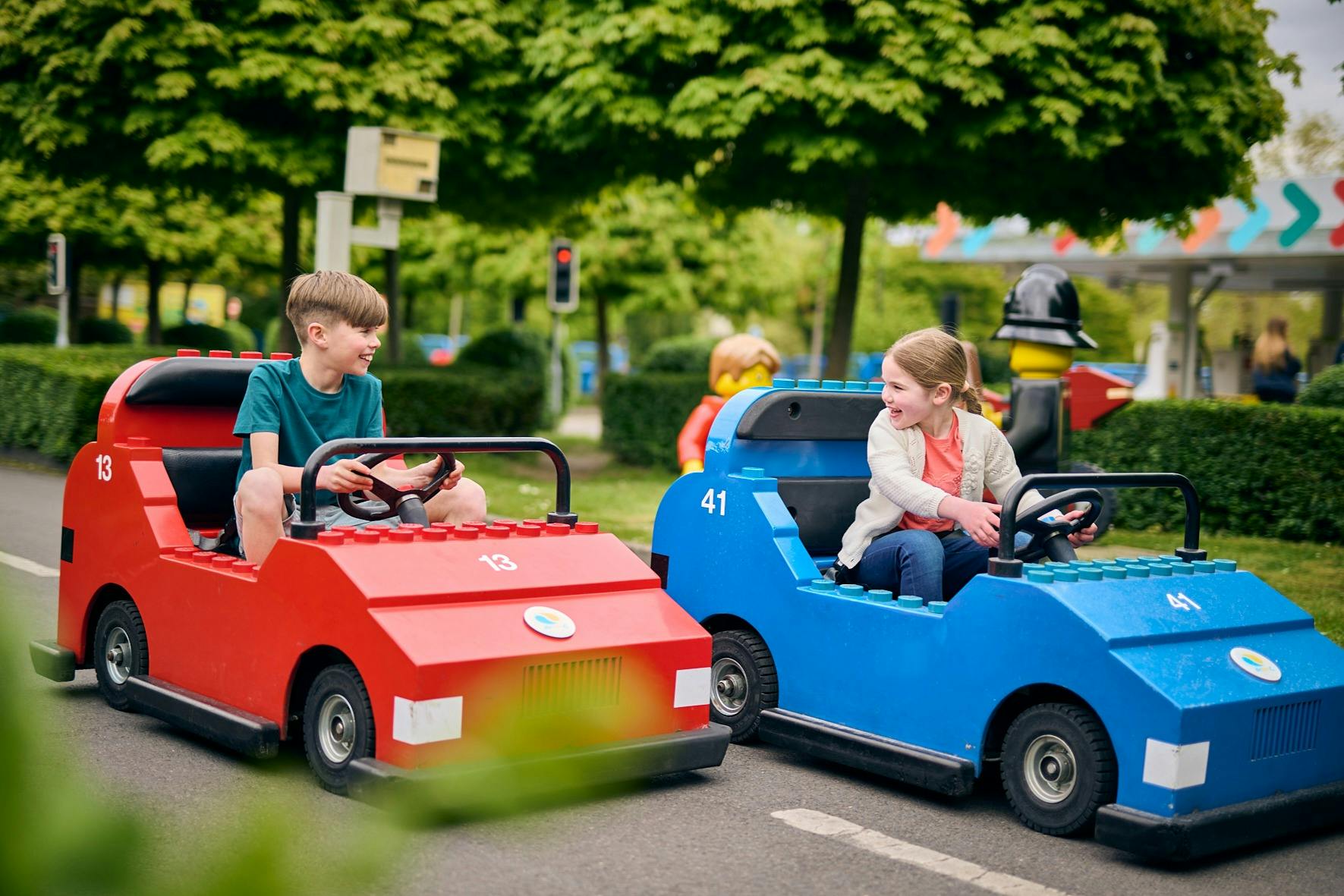 Uma criança a conduzir um carro de brincar azul num parque, sorrindo e olhando para outra criança num carro de brincar vermelho. Árvores e arbustos rodeiam-nos.