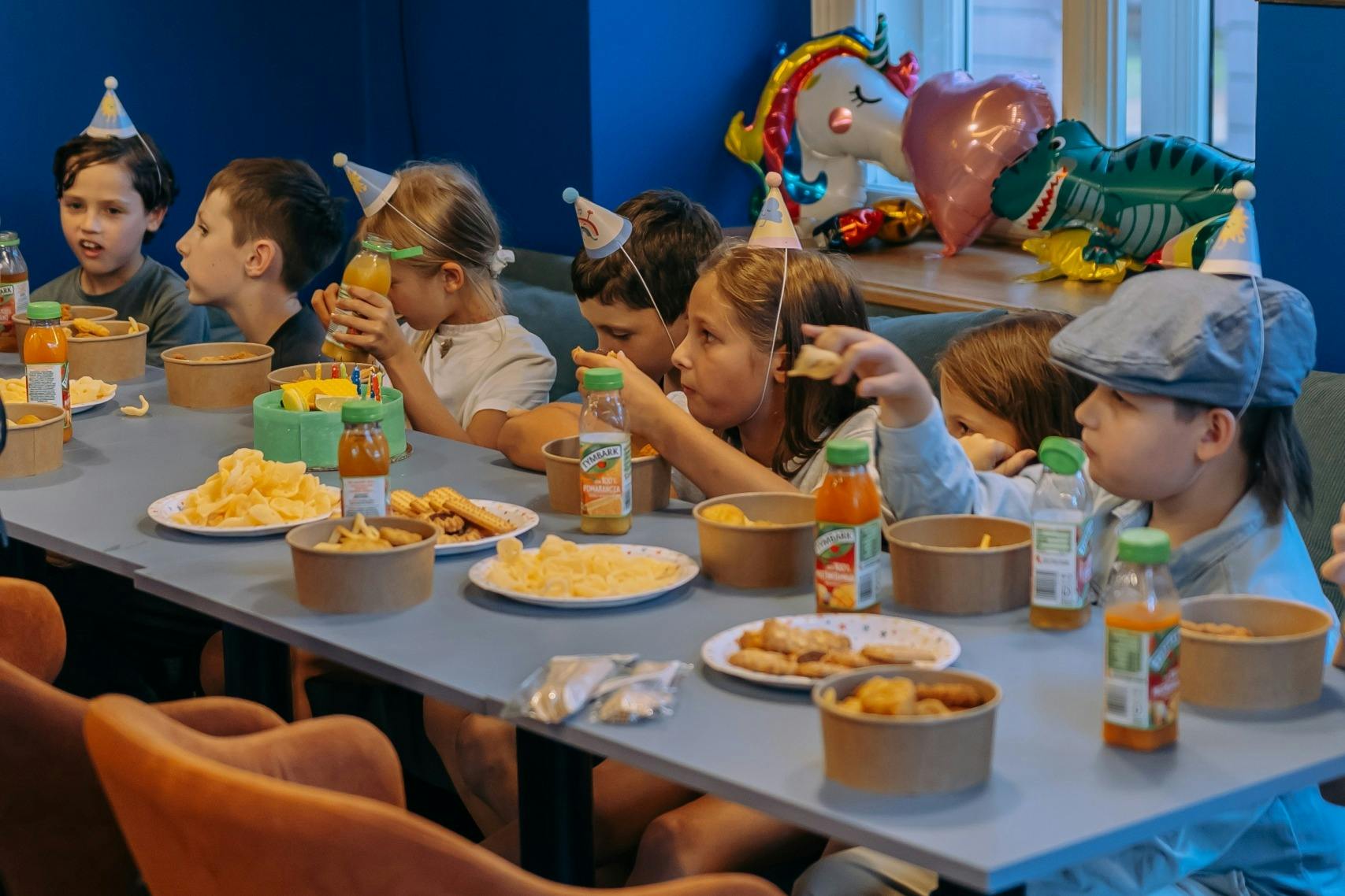 Children seated at a table with snacks and drinks, wearing party hats, in a festive setting with a balloon decoration.