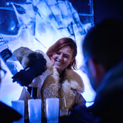 En kvinna i en tjock pälsfodrad kappa häller en drink i isglas och ler mot två sponsorer i en svagt upplyst, blåtonad miljö.