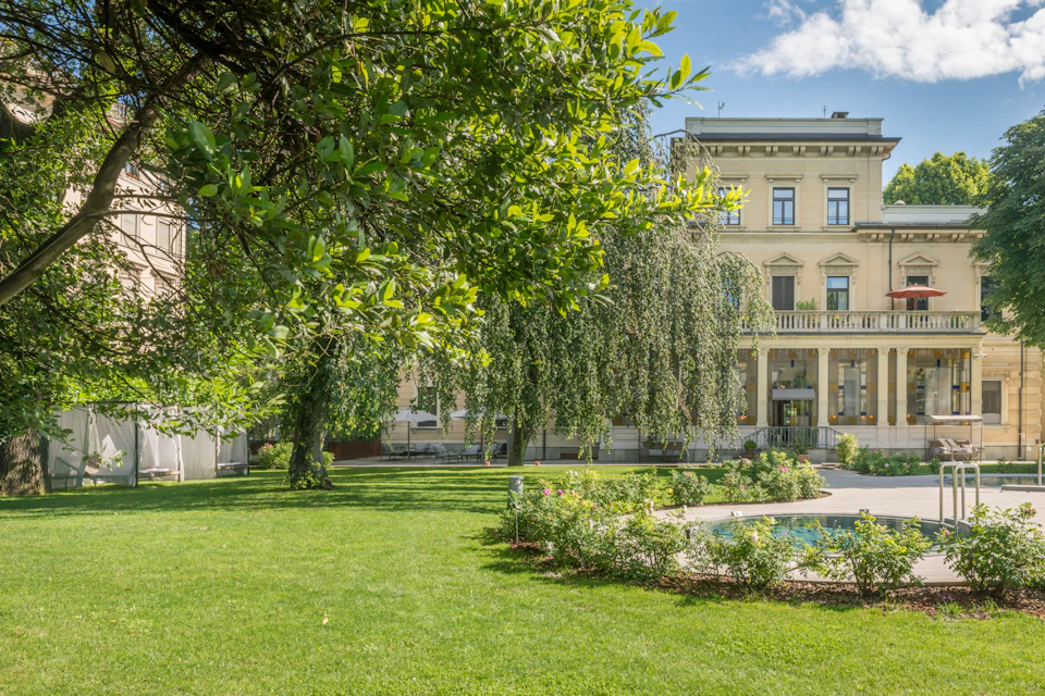 A lush garden with green grass, trees, and a fountain, in front of a classic building with a beige facade under a blue sky.