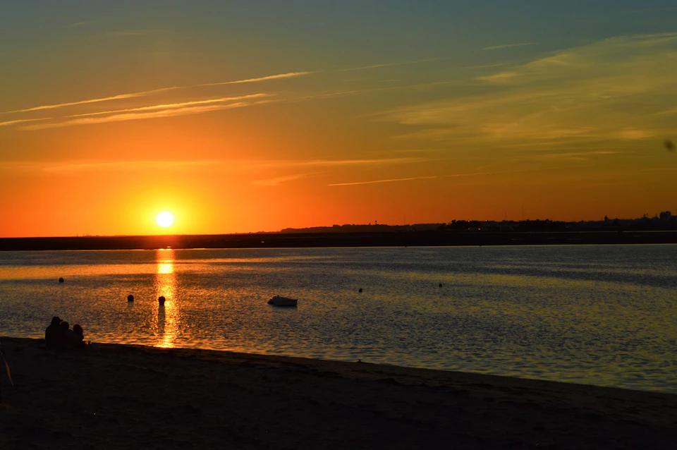 Sunset over calm water with a few buoys and a small boat. Sandy shoreline in the foreground, clear sky with thin clouds.