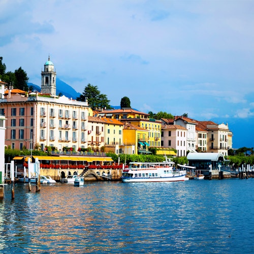 Colorful buildings and a dock with boats along a waterfront under a partly cloudy sky, with a tower visible in the background.