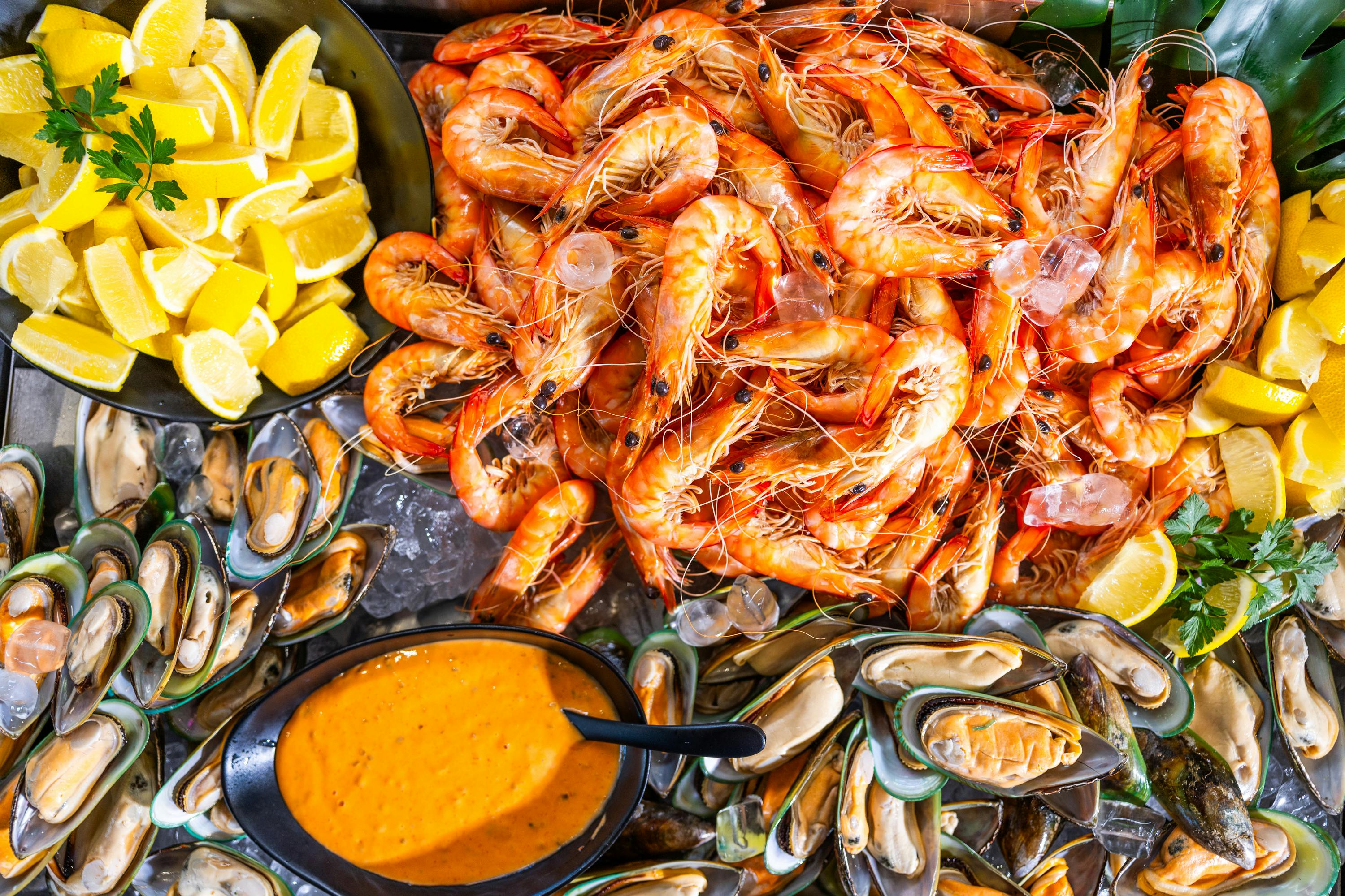 Μια ποικιλία από φρέσκα θαλασσινά, όπως γαρίδες, μύδια και φέτες λεμονιού, τοποθετημένα γύρω από ένα μπολ με σάλτσα πορτοκαλιού.