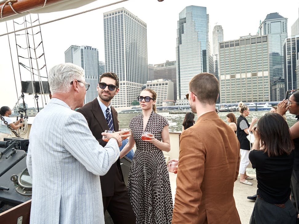 A group of people in formal attire conversing on a boat with a city skyline and water in the background.