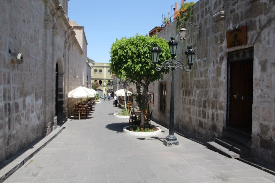 Cobblestone street with outdoor tables and umbrellas, flanked by old stone buildings. A tree and streetlamp are centered.