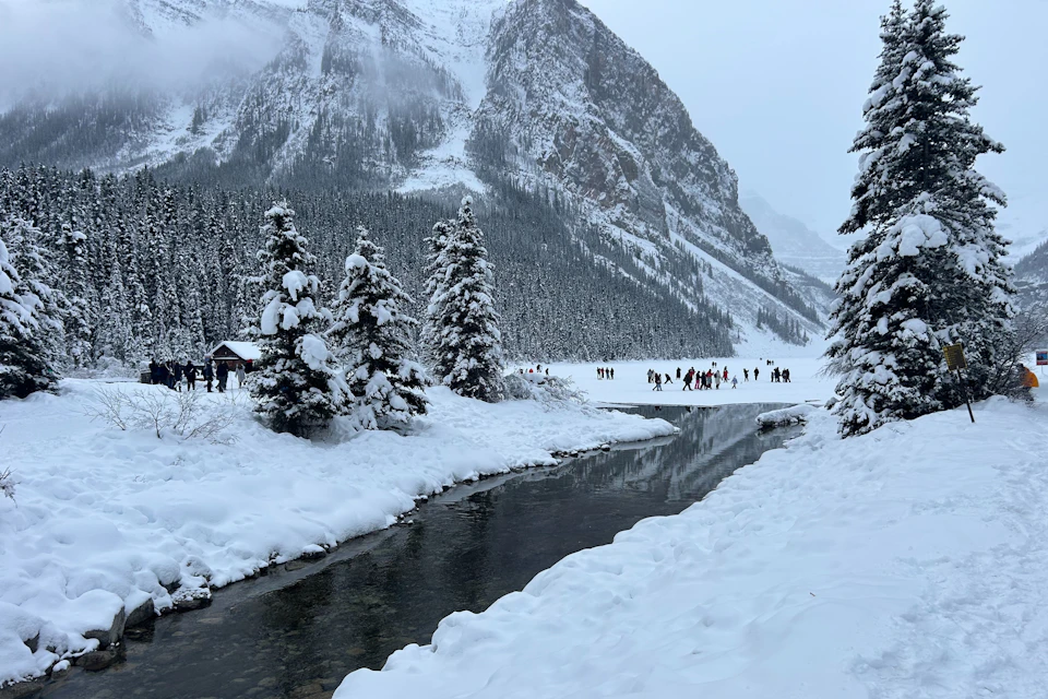 Snow-covered landscape with a river, tall pine trees, and distant people on a frozen lake. A mountain range is in the background.