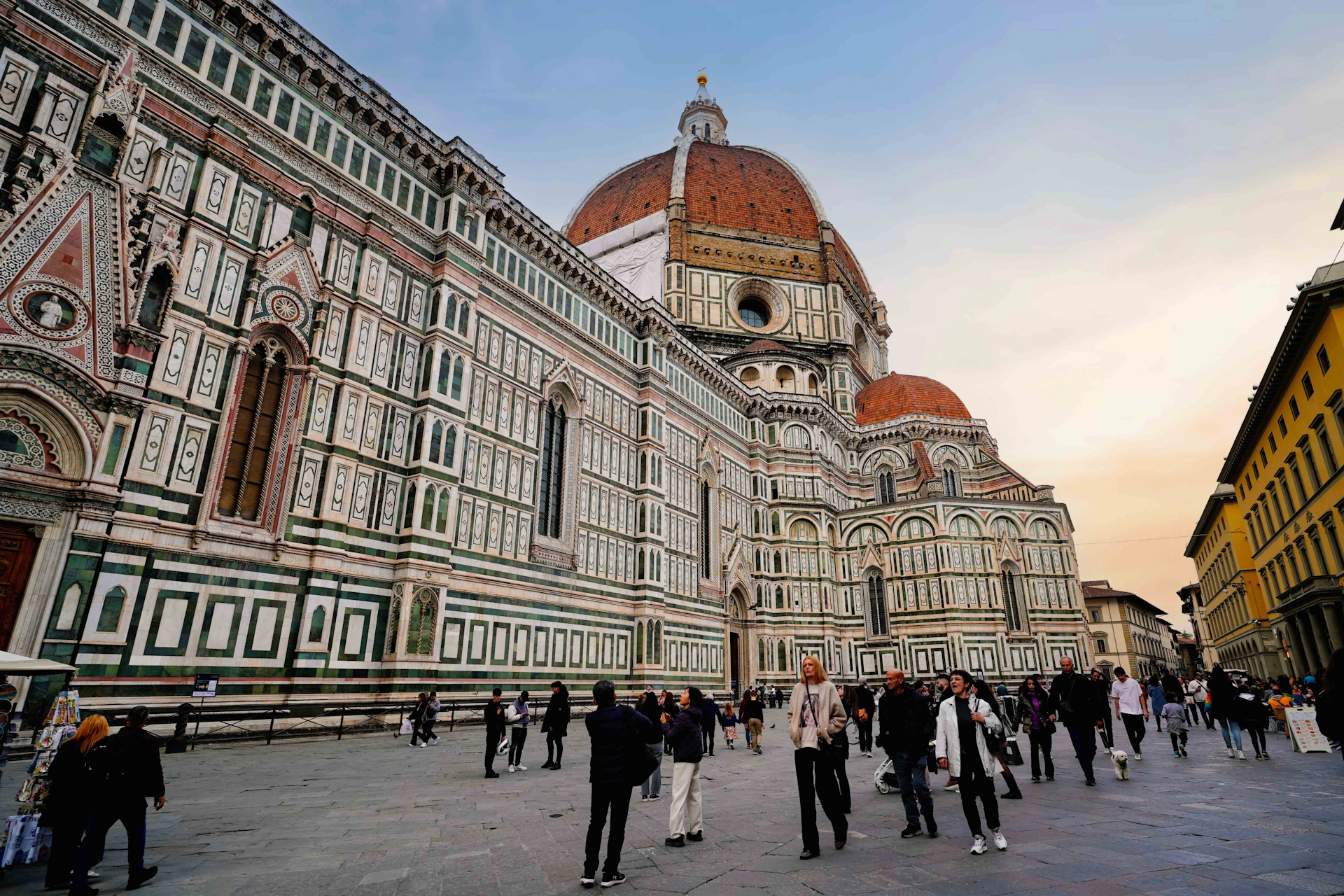 People walking in front of the Florence Cathedral, featuring its intricate facade and large dome under a clear sky.