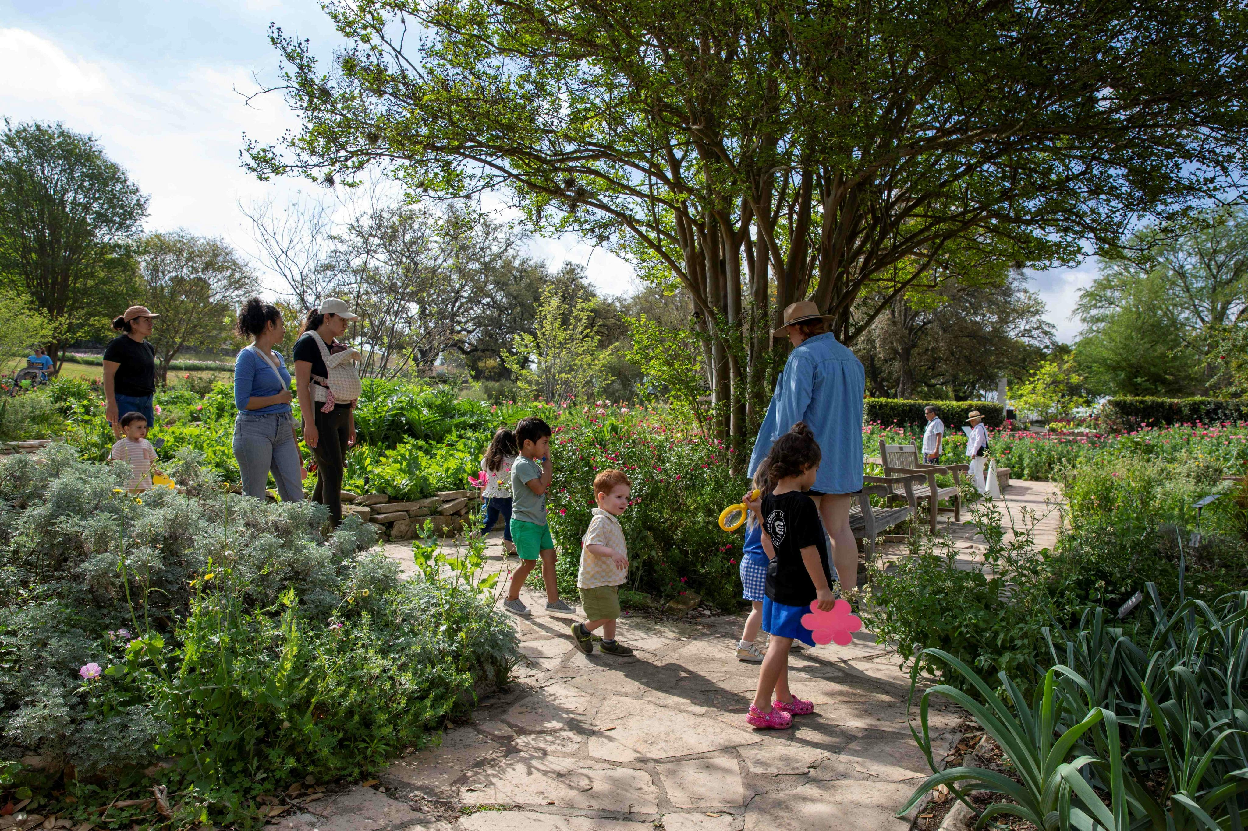 Nens i adults caminant per un camí amb jardí envoltat de vegetació i arbres en un dia assolellat.