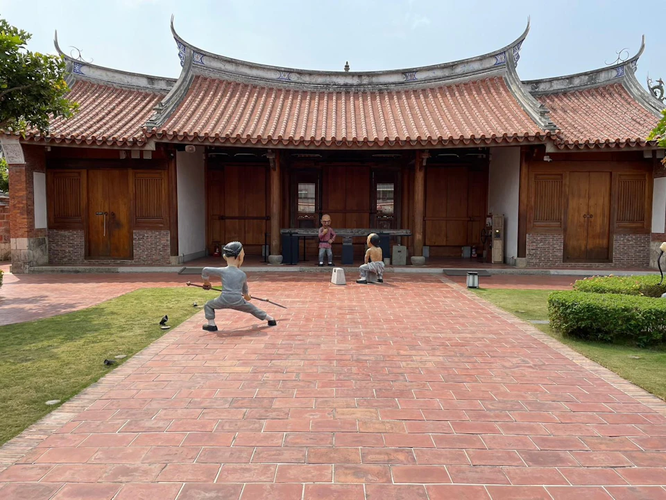 A courtyard with a traditional building, sculptures of two figures in martial poses, and one seated figure in the center.