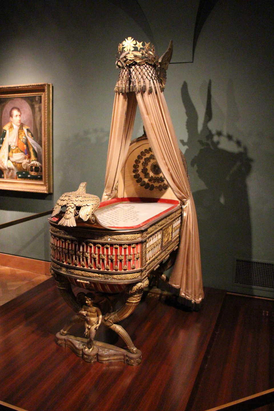Ornate crib with draped canopy and eagle decoration in a dimly lit room. Portrait of a man in military uniform on the wall.