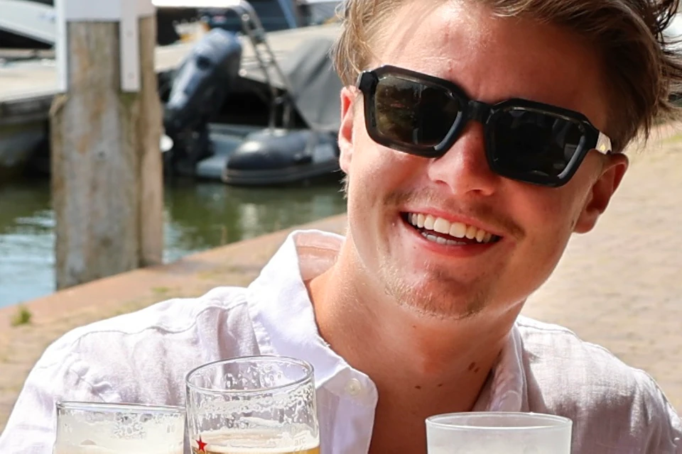 Two people sitting at an outdoor table near a harbor, smiling and toasting with glasses of beer on a sunny day.