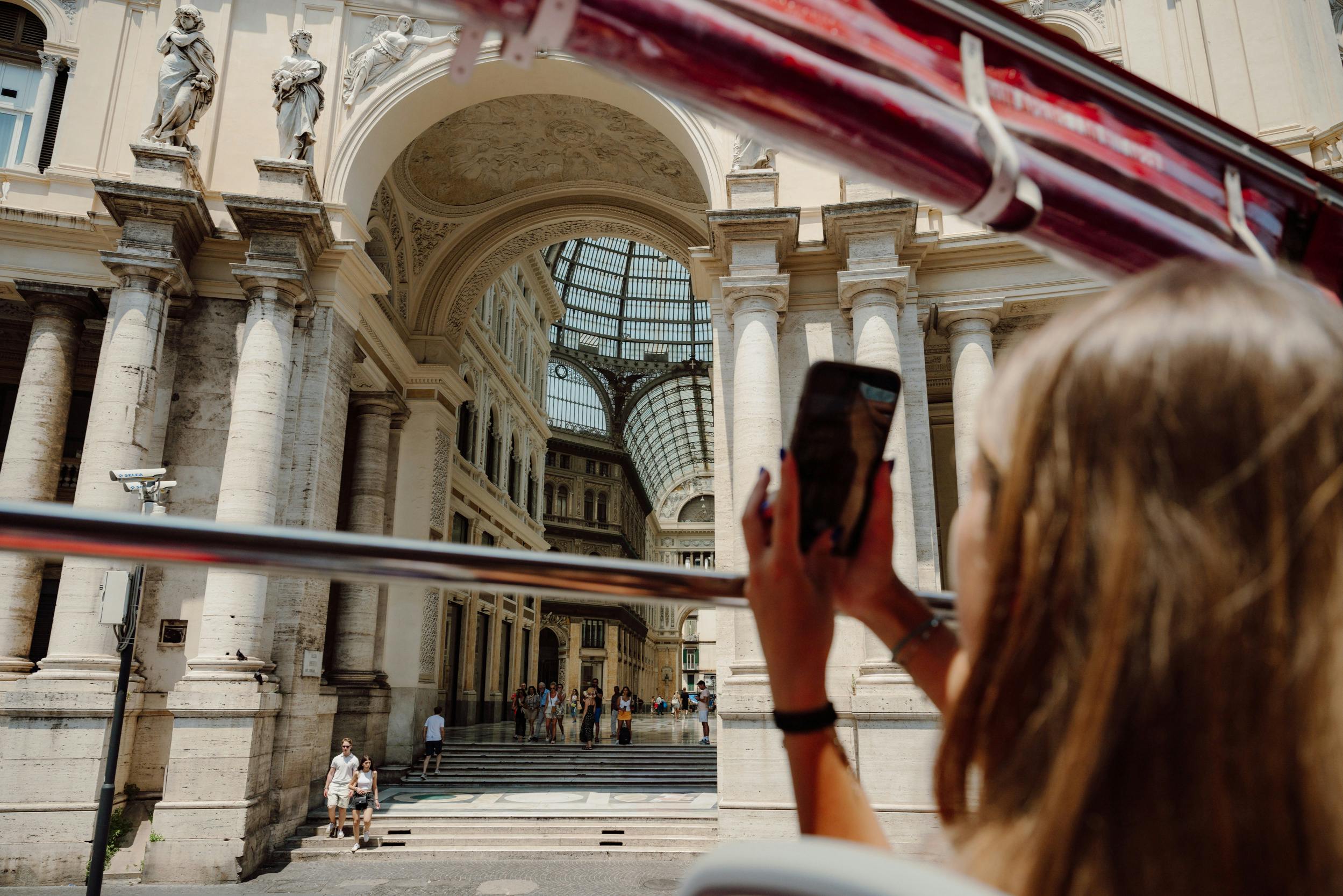 Una persona con in mano uno smartphone scatta una foto di un edificio decorato con archi e soffitto di vetro, visto da un autobus.