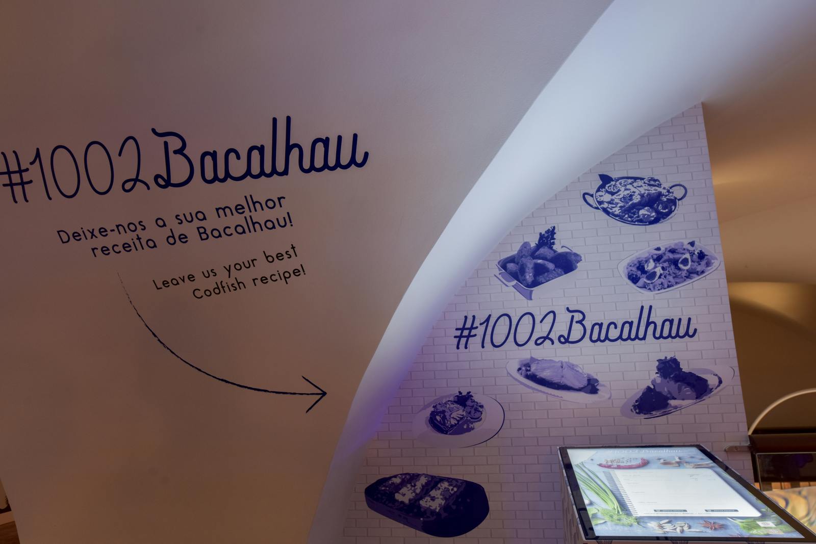 Curved white wall with &#34;Bacalhau&#34; text, food images, hashtags, and &#34;Leave us your best Codfish recipe!&#34; invitation.