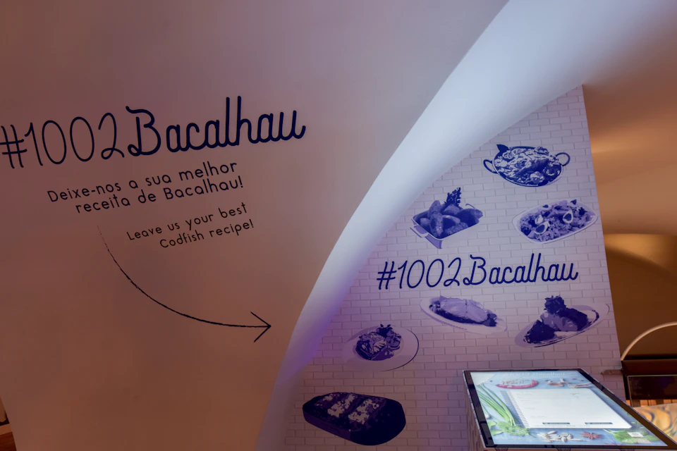 Curved white wall with "Bacalhau" text, food images, hashtags, and "Leave us your best Codfish recipe!" invitation.