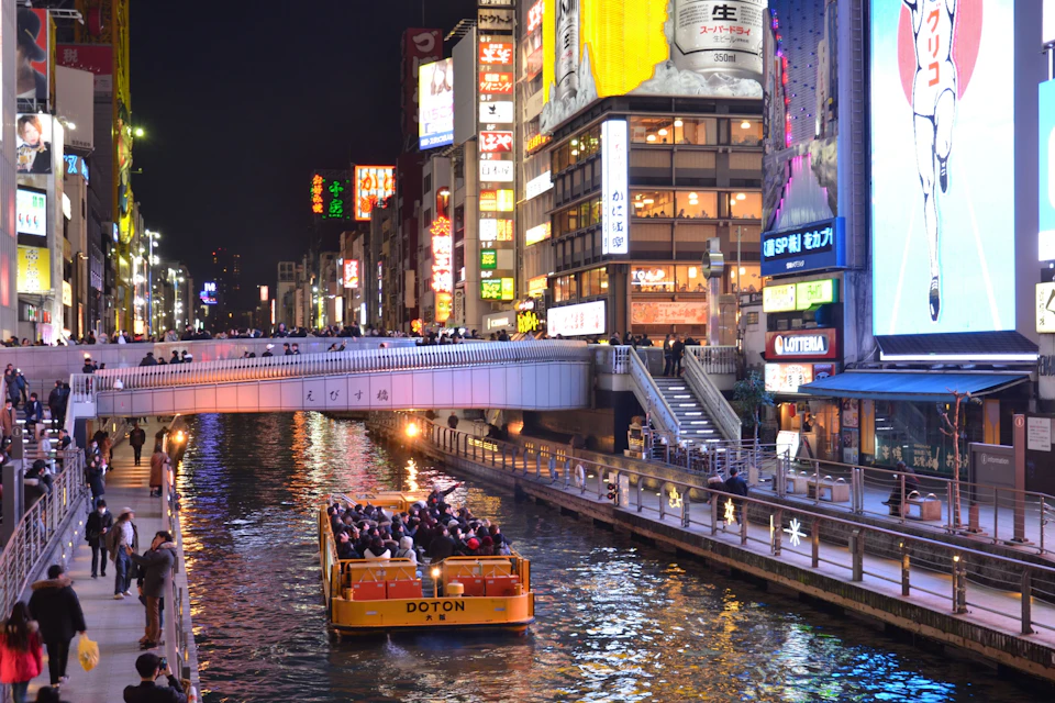 A brightly lit canal at night with a yellow boat carrying passengers, surrounded by illuminated buildings and a bridge.