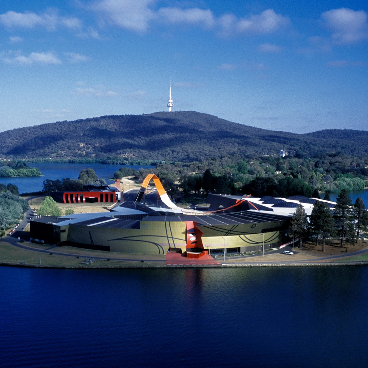 Aerial view of a modern building with artistic design near a lake, surrounded by greenery, with hills and a tower in the distance.
