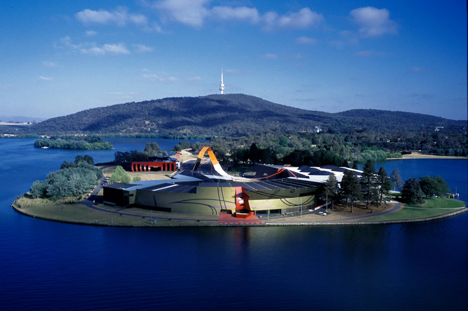 Aerial view of a modern building with artistic design near a lake, surrounded by greenery, with hills and a tower in the distance.