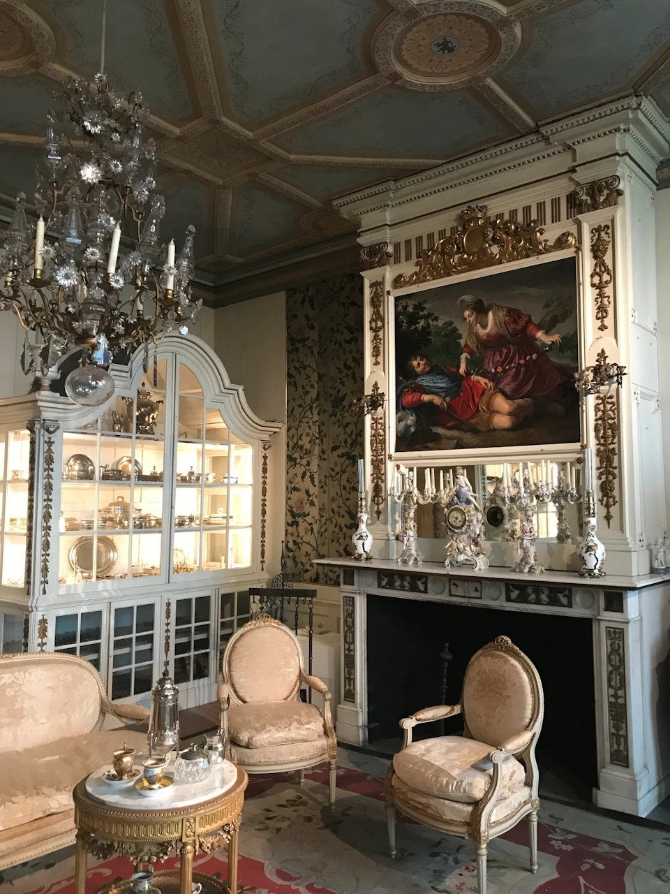 Elegant room with ornate chandelier, antique furniture, and glass display cabinet. Large painting and decorative pieces above a fireplace.
