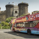 A colorful sightseeing bus in Naples in front of an ancient castle with large round towers.