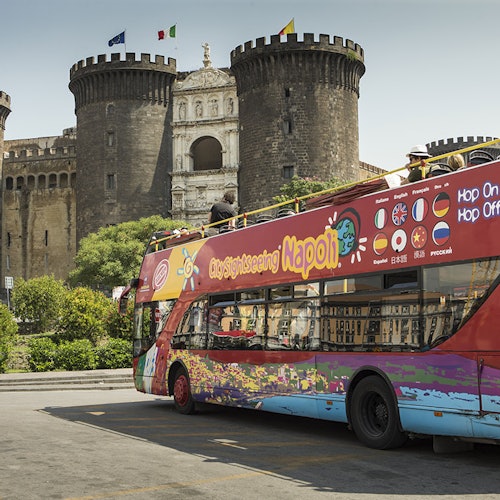 A colorful sightseeing bus in Naples in front of an ancient castle with large round towers.