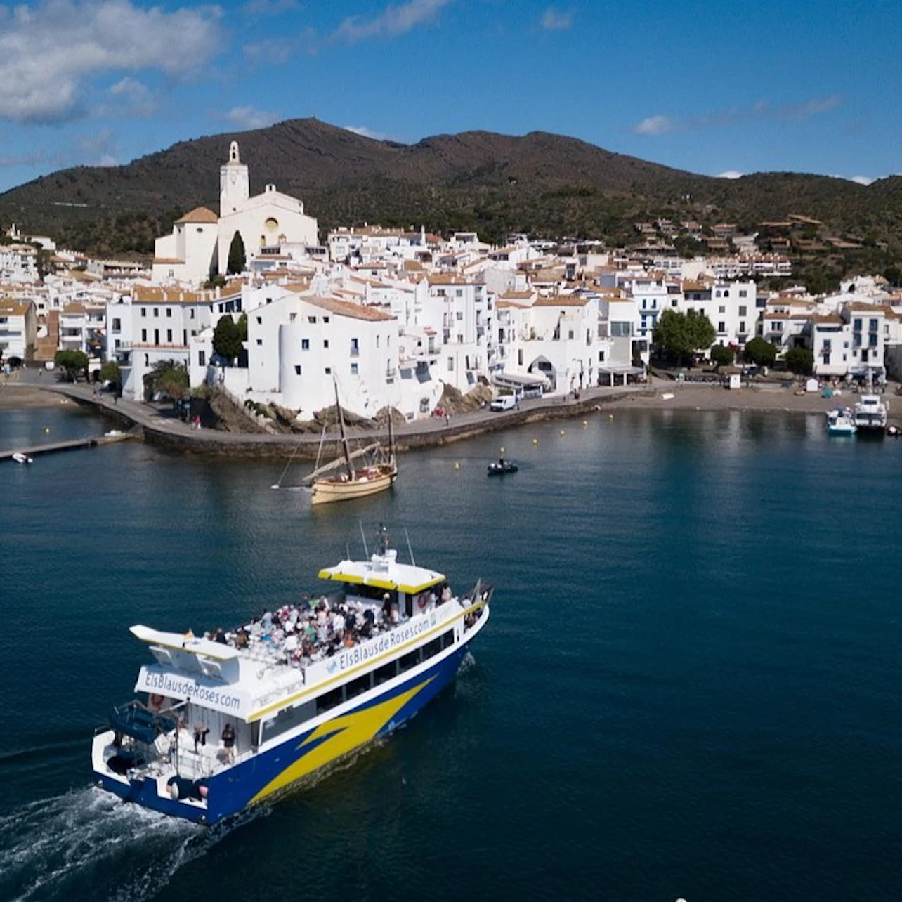 A boat filled with people sails near a coastal village with white buildings, a church, and hills in the background under a blue sky.