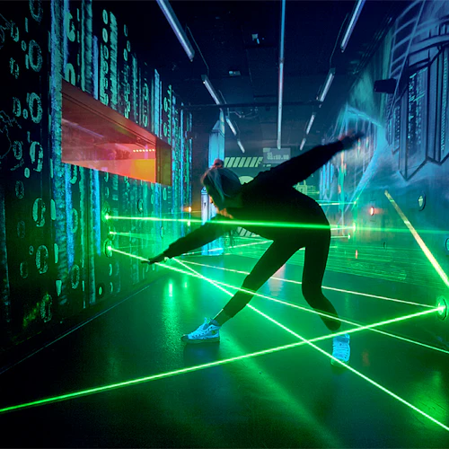En person i ett mörkt rum navigerar genom gröna laserstrålar, med digital grafik och neonljus på väggarna.