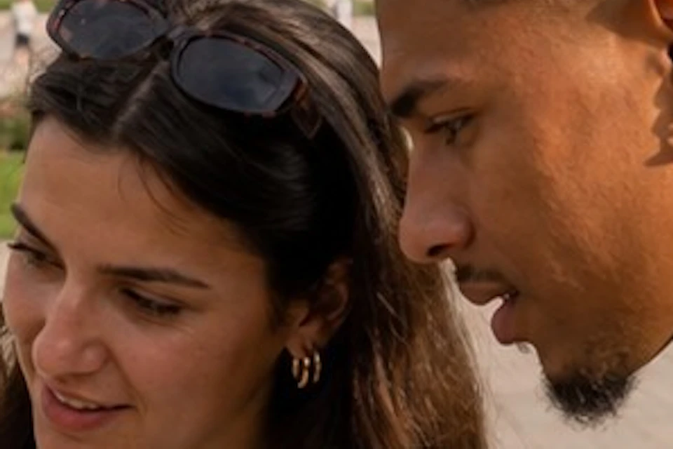 Two people outdoors looking at a smartphone, with a column and historic buildings in the background on a sunny day.